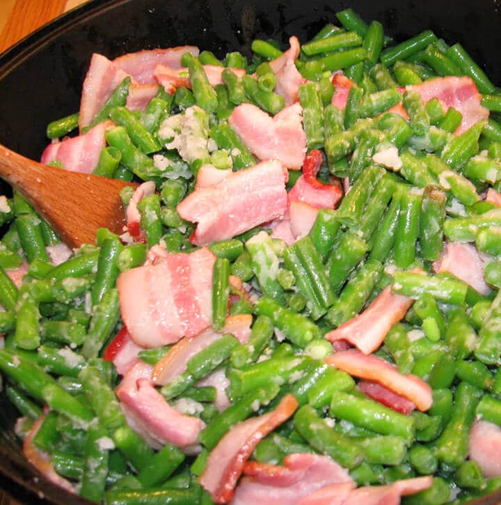 Stirring green beans and bacon with a spoon in a large pot.