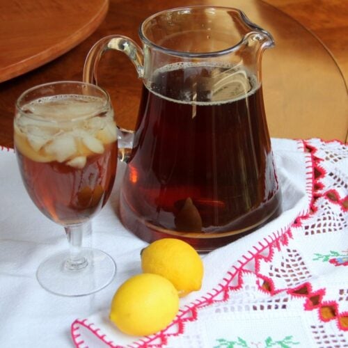A glass and a pitcher of Southern Style sweet tea with lemon slices sit on a white embroidered cloth, accompanied by two whole lemons.