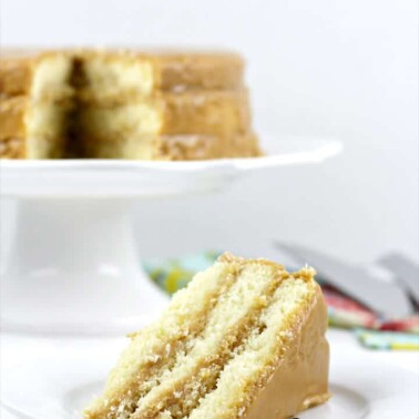 Slice of caramel cake on a plate with whole cake in background.