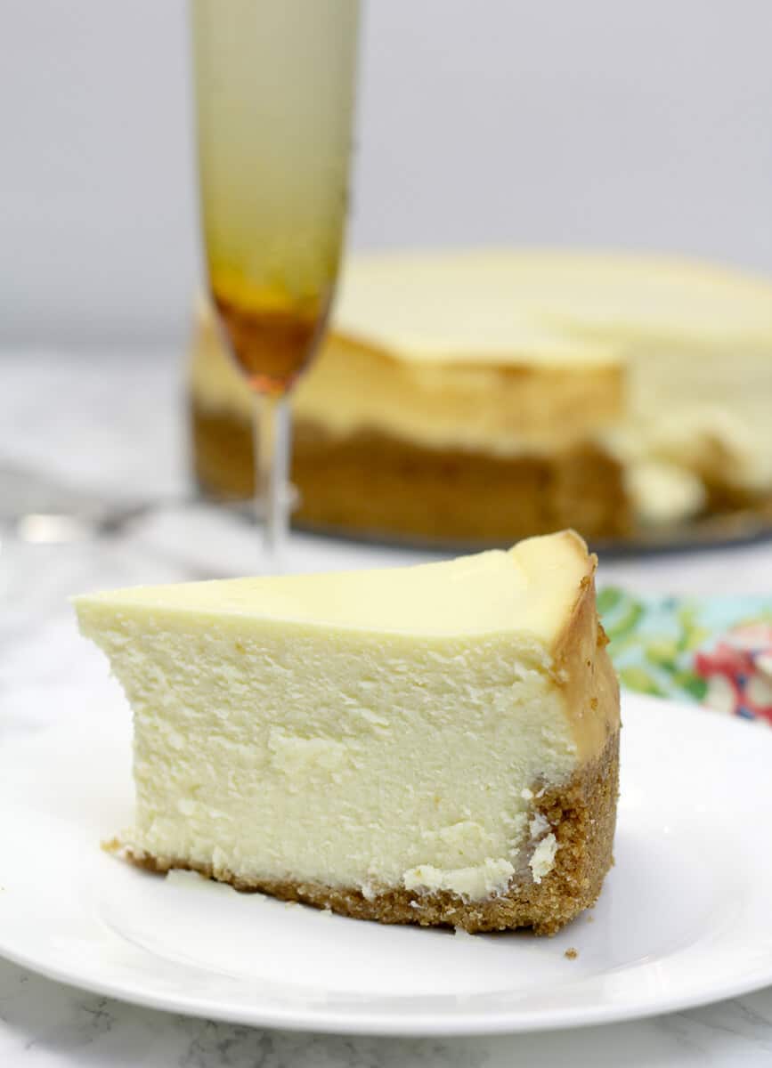 Side view of a slice of plain cheesecake on a plate with a champagne glass in the background.