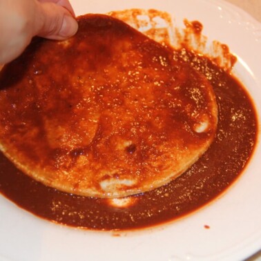 A hand dipping a tortilla in enchilada sauce.