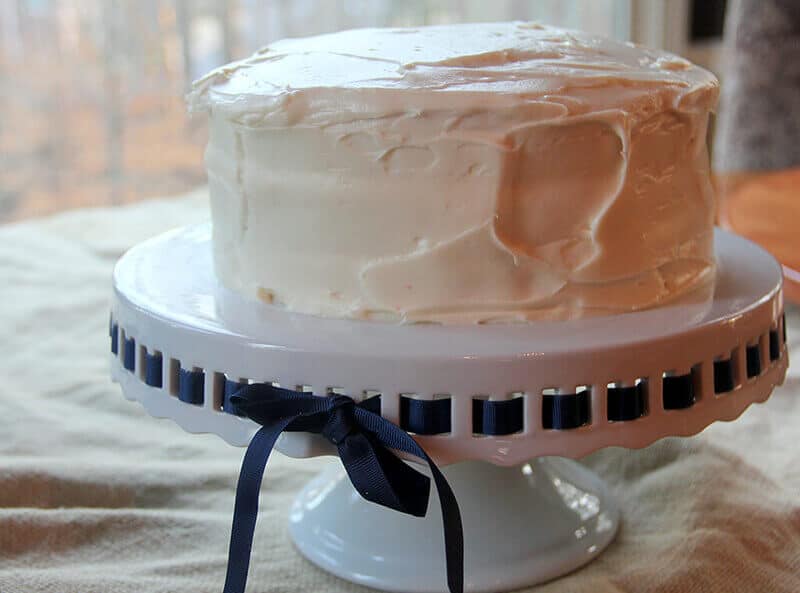 A whole frosted Red Velvet Cake on a cake stand with a blue ribbon woven through the sides of the stand. 