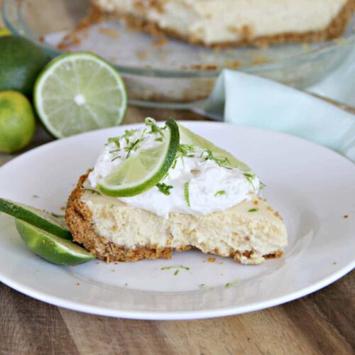 A slice of key lime pie topped with whipped cream and lime slices sits on a white plate, showcasing the vibrant flavors of this classic key lime pie, with whole and cut limes in the background.