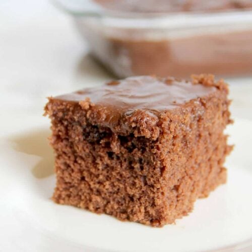 A square piece of chocolate Coca-Cola cake with rich chocolate frosting sits on a white plate, while the rest of the cake rests in a glass dish in the background.