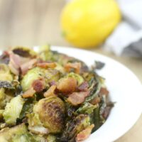 Serving of sauteed brussels sprouts in a white dish with bacon and a lemon.
