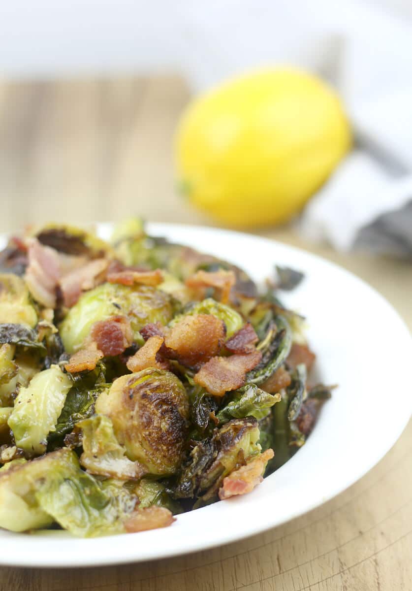 Serving of sauteed brussels sprouts in a white dish with bacon and a lemon.