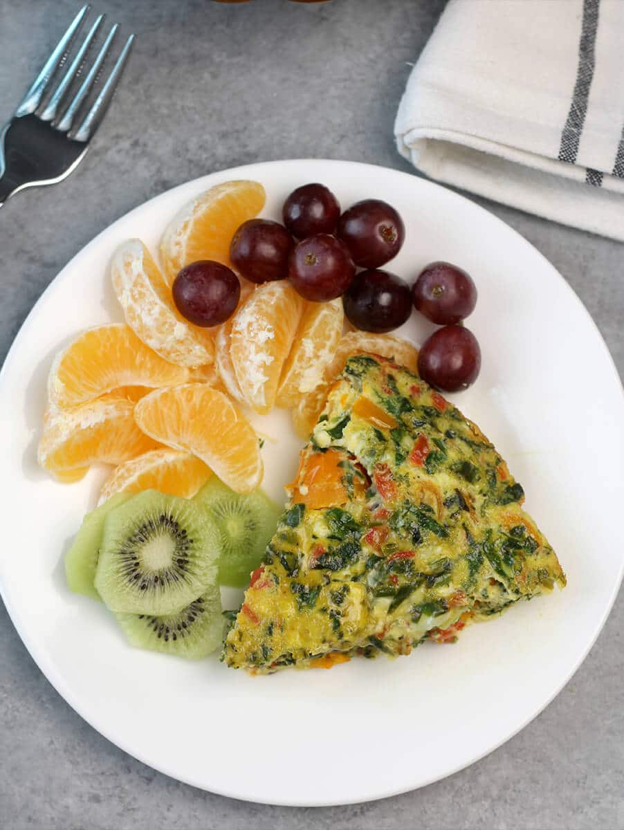 Overhead view of slice of easy crustless quiche on a plate with grapes, tangerine slices, and kiwi. 