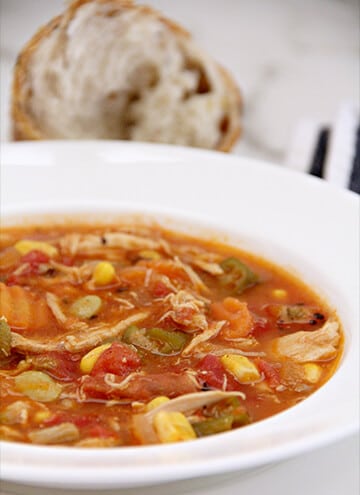 A bowl of chicken vegetable soup with bread in the background.