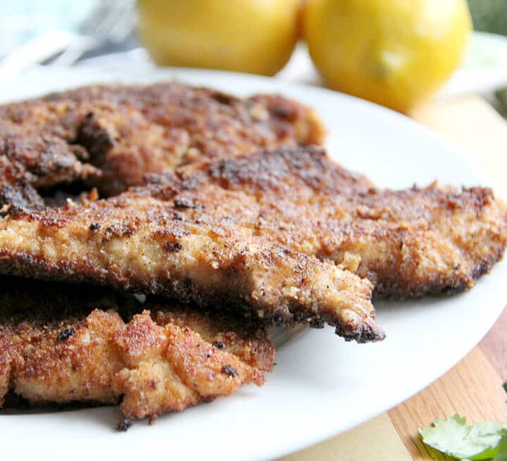 White platter of parmesan chicken cutlets with lemons in the background for an entree for a romantic dinner at home.
