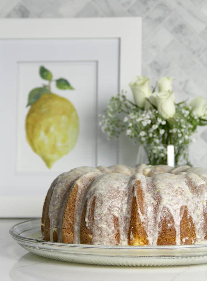 A pound cake drizzled with lemon glaze icing with a vase of flowers in the background.