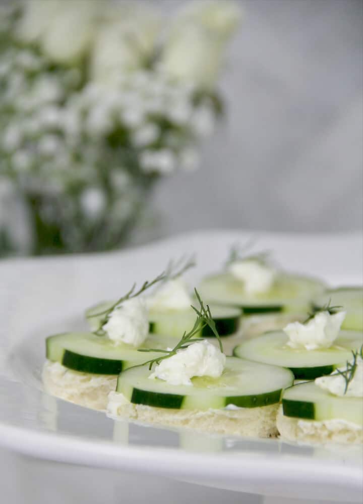 Photo of a platter of cucumber sandwiches with a vase of flowers in the background.