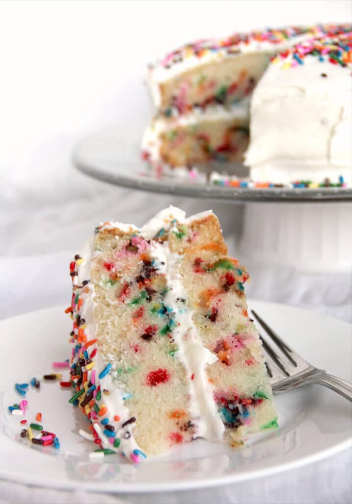 Slice of homemade funfetti cake on a white plate with a whole cake in the background.