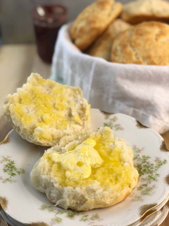 Buttered buttermilk biscuit on a plate with bowl of biscuits in background.