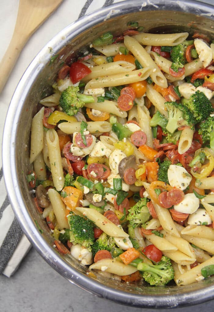 A bowl of pasta salad with pepperoni and other pizza toppings with a wooden spoon on the side.