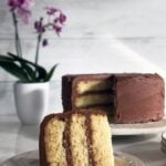 A slice of moist yellow cake on a plate with the whole cake in the background with chocolate frosting.