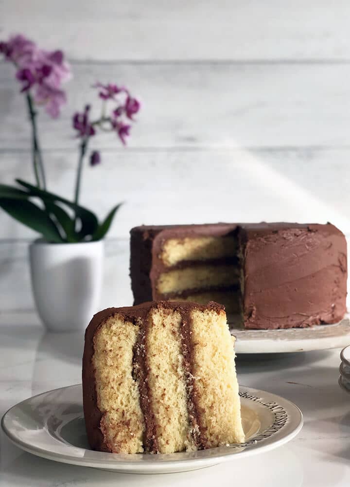 A slice of moist yellow cake on a plate with the whole cake in the background with chocolate frosting.