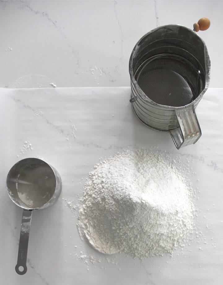 Measuring cup and flour sifter with sifted flour on parchment paper.