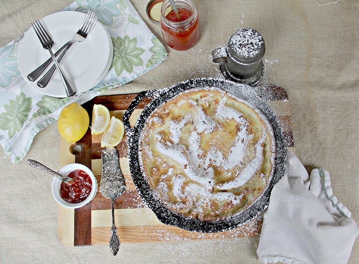 Overhead photo of a skillet of Dutch baby german pancake with plates, lemons, and jelly on the side.