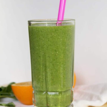 A glass of super green smoothie with a pink straw and an orange on a cutting board.