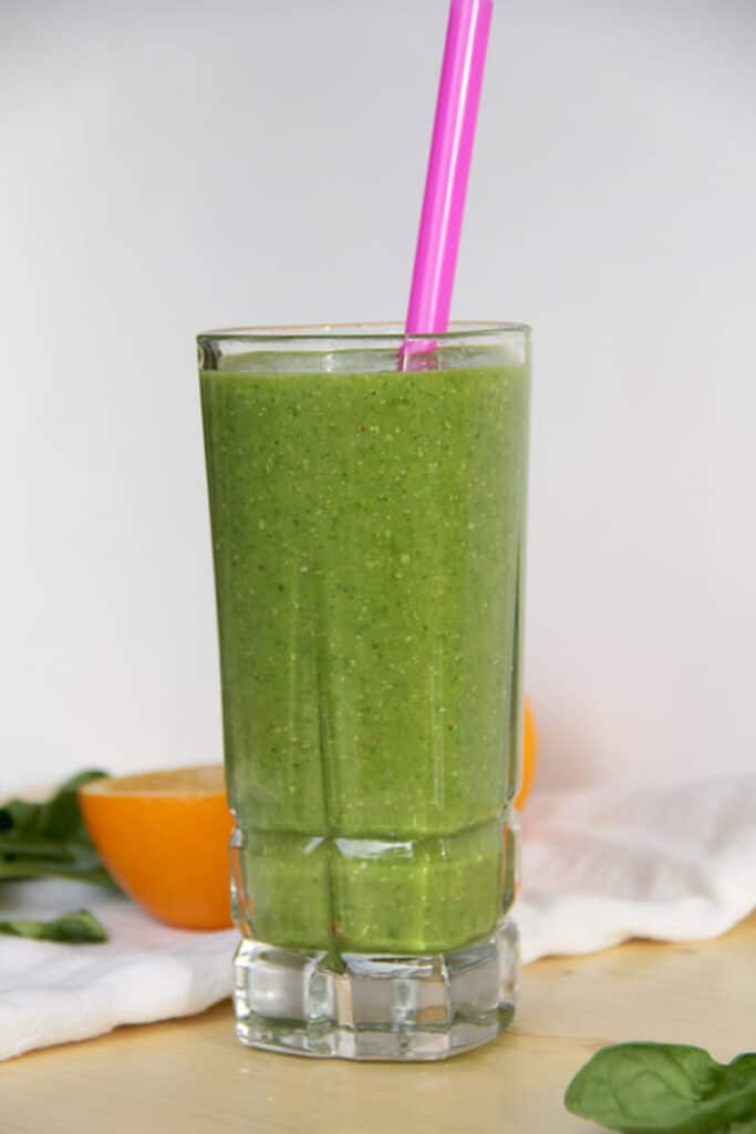 A glass of super green smoothie with a pink straw and an orange on a cutting board.