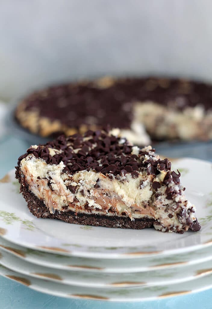 A slice of chocolate chip cheesecake on a stack of plates with the whole pie in the background.