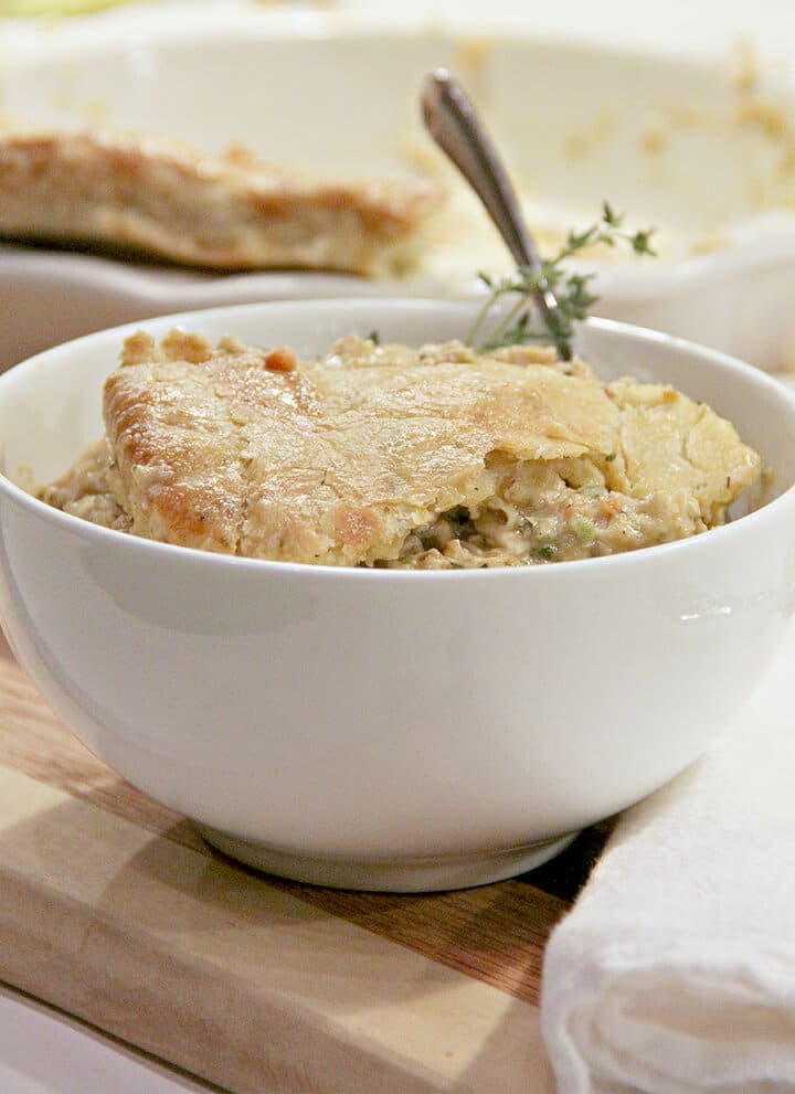 Serving of double crust chicken pot pie in a white bowl with a sprig of thyme on a wooden board.