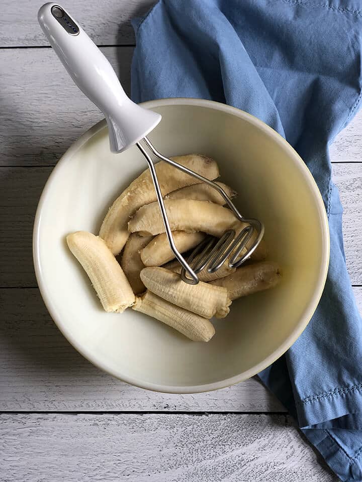Bananas in a bowl with a masher next to a blue towel.