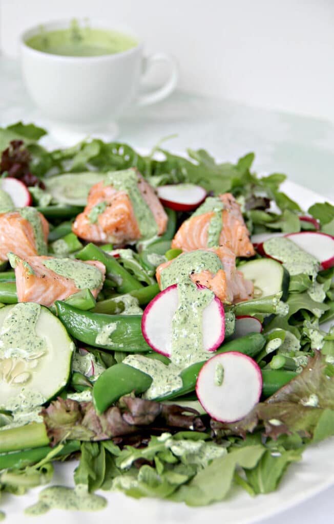 Side angle of lettuce and arugula with fresh spring vegetables and baked salmon on a white plate with a cup of dressing in the background.