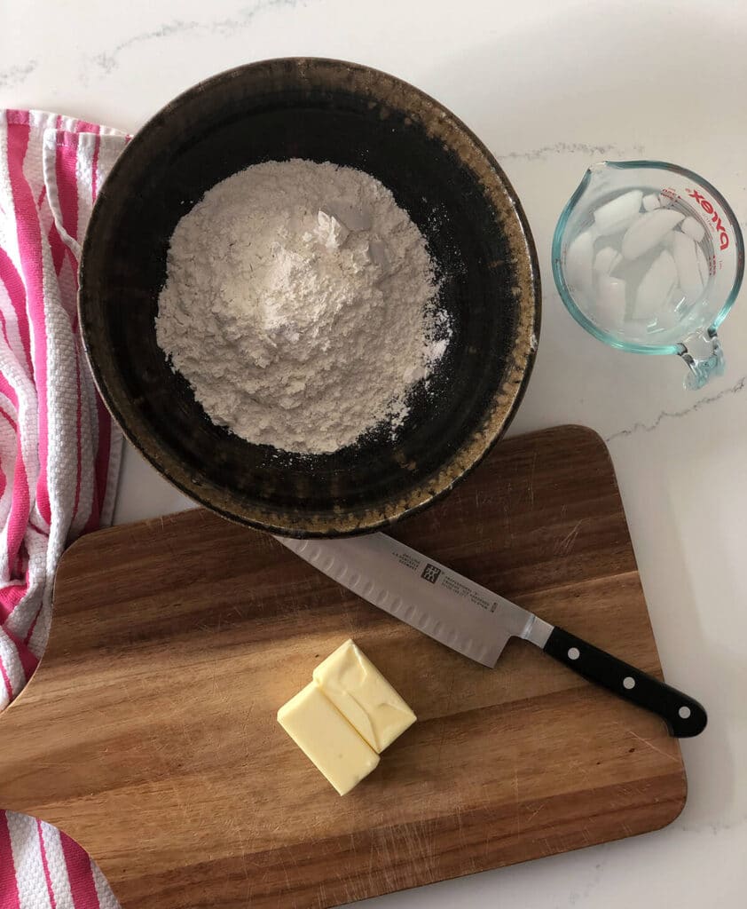 A bowl of flour and chunks of butter on a cutting board next to a measuring cup of ice water.