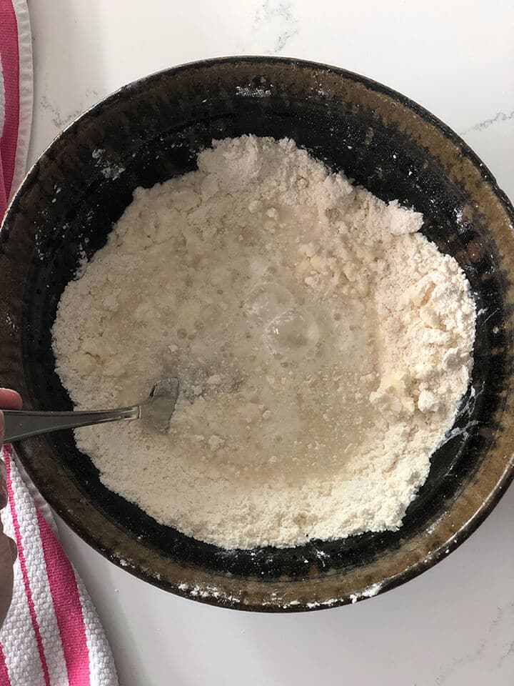 A brown bowl with flour and ice water with a fork stirring to make chicken and dumplings.