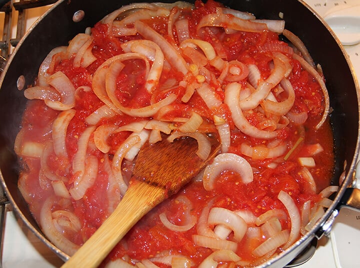 A wooden spoon stirring onions in a skillet for Mediterranean fish.