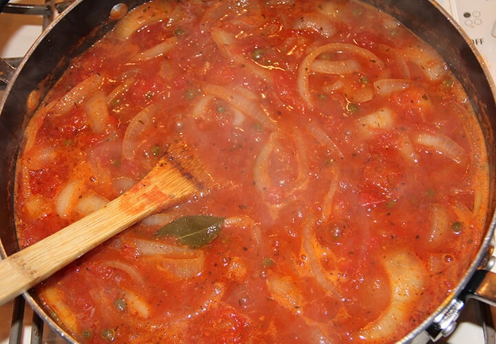 Onions and tomatoes simmering in a large skillet with a wooden spoon.