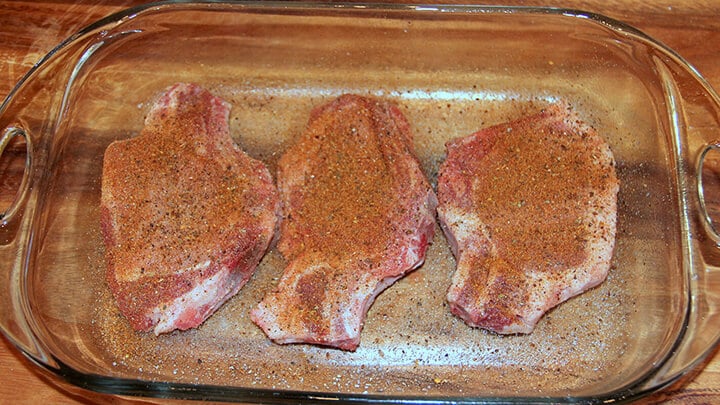 Three raw pork chops with seasoning in a glass baking dish.