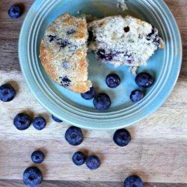 A blueberry muffin on a blue plate on a cutting board with blueberries.