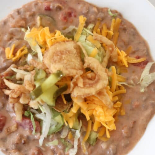 A bowl of cheeseburger soup with cheese and pickles toppings.
