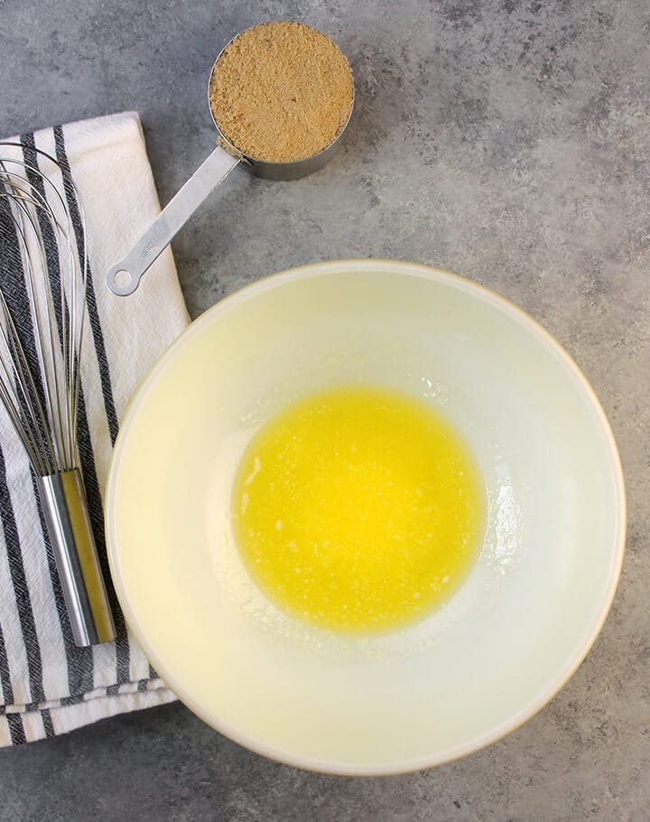 A bowl with melted butter with a cup of brown sugar and a whisk on the side.