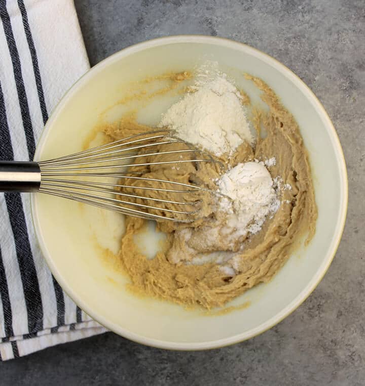 A wire whisk mixing together eggs, brown sugar, and flour in a bowl on a grey counter.