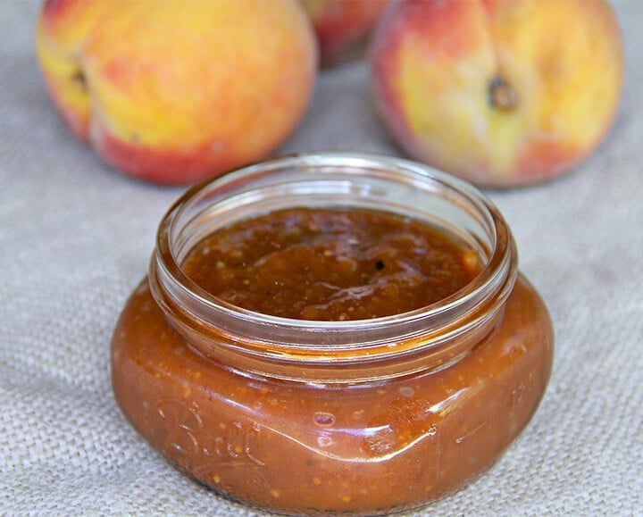A small jar of peach bbq sauce on burlap with peaches in the background.
