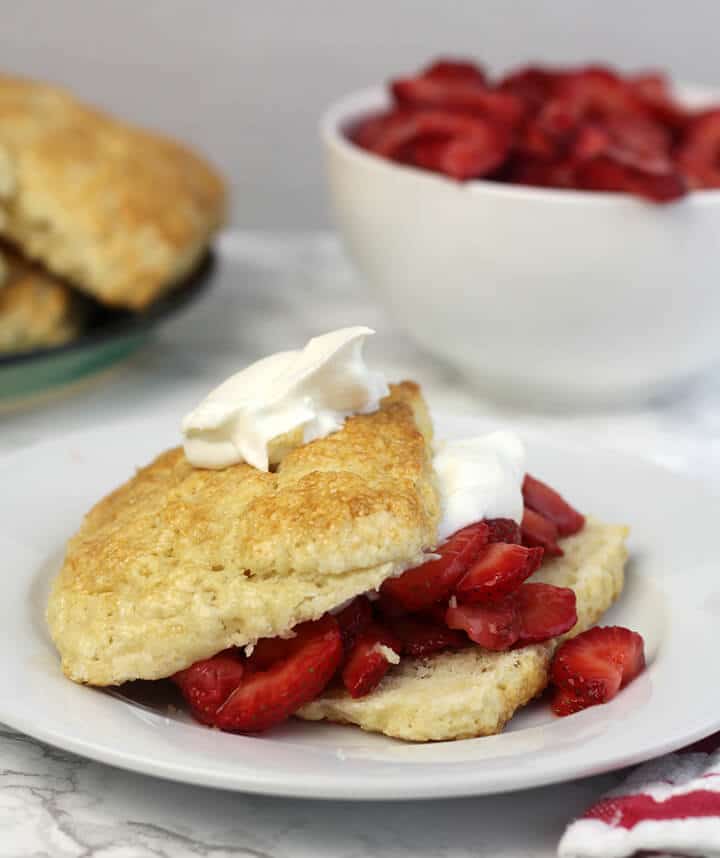 Strawberry shortcake on a white plate with a bowl of strawberries in the background.