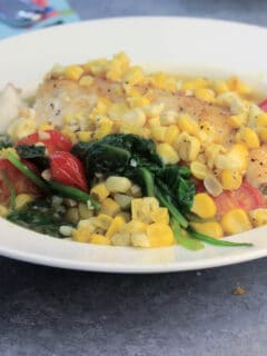 Oven baked fish in a white bowl with fresh summer vegetables like spinach, corn, and tomatoes will be one of your weeknight favorites! This recipe is quick and easy and everyone loves it!