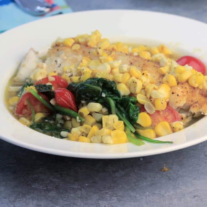 Oven baked fish in a white bowl with fresh summer vegetables like spinach, corn, and tomatoes will be one of your weeknight favorites! This recipe is quick and easy and everyone loves it!