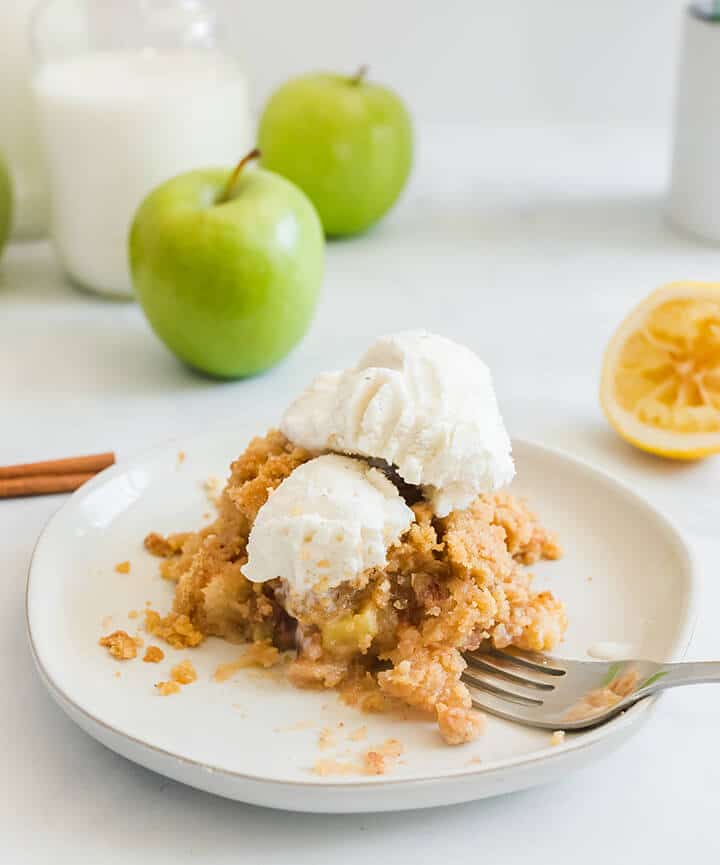 A serving of apple crisp on a white plate with apples in the background.