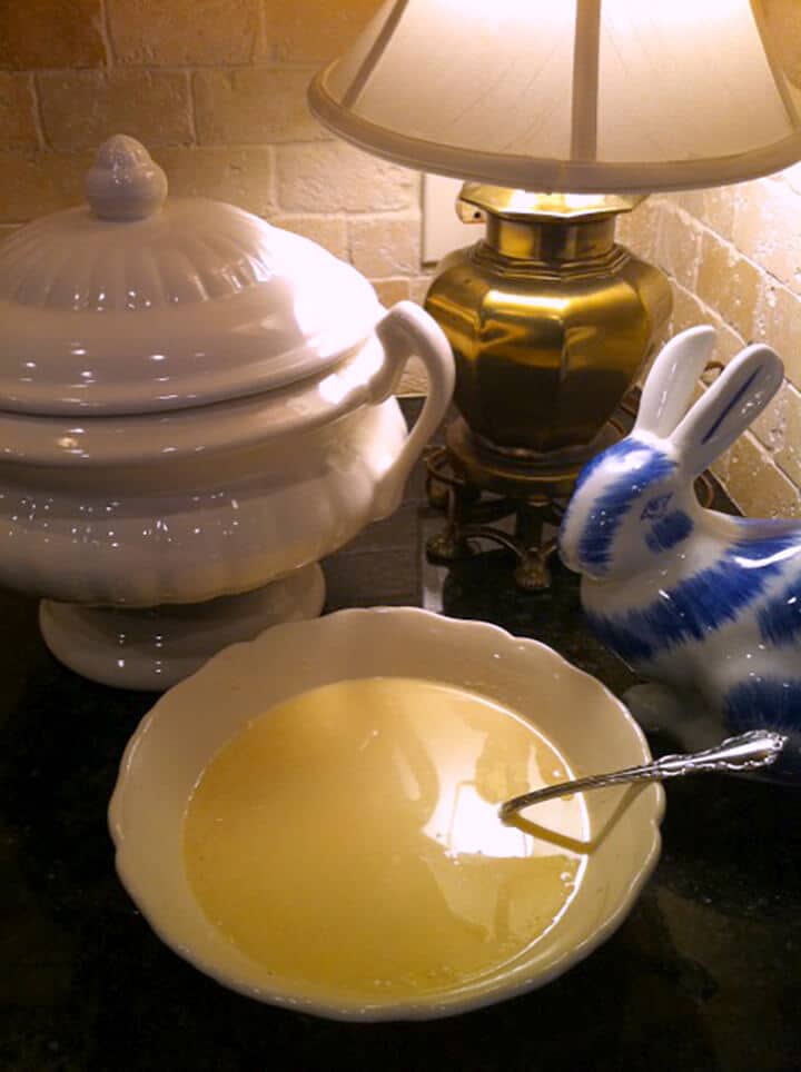 A bowl of Grandma's Chicken Soup on a table with a white and blue ceramic rabbit and a lamp.