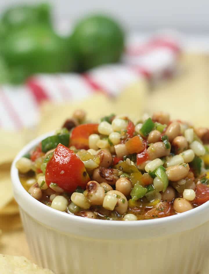 A white bowl filled with texas caviar and surrounded by chips with a red striped towel in the background.