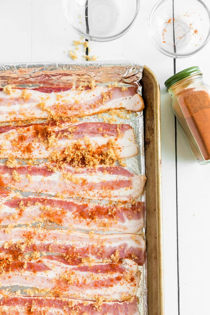 Raw bacon slices sprinkled with brown sugar and cayenne pepper on a sheet pan.