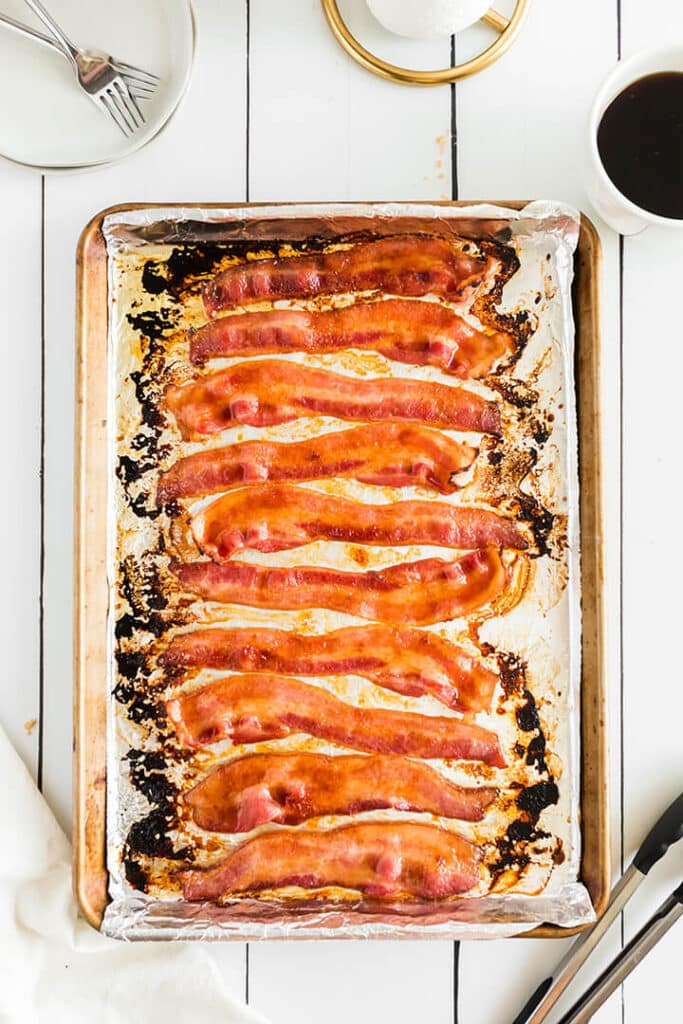 A sheet pan of oven baked bacon with tongs on the side.