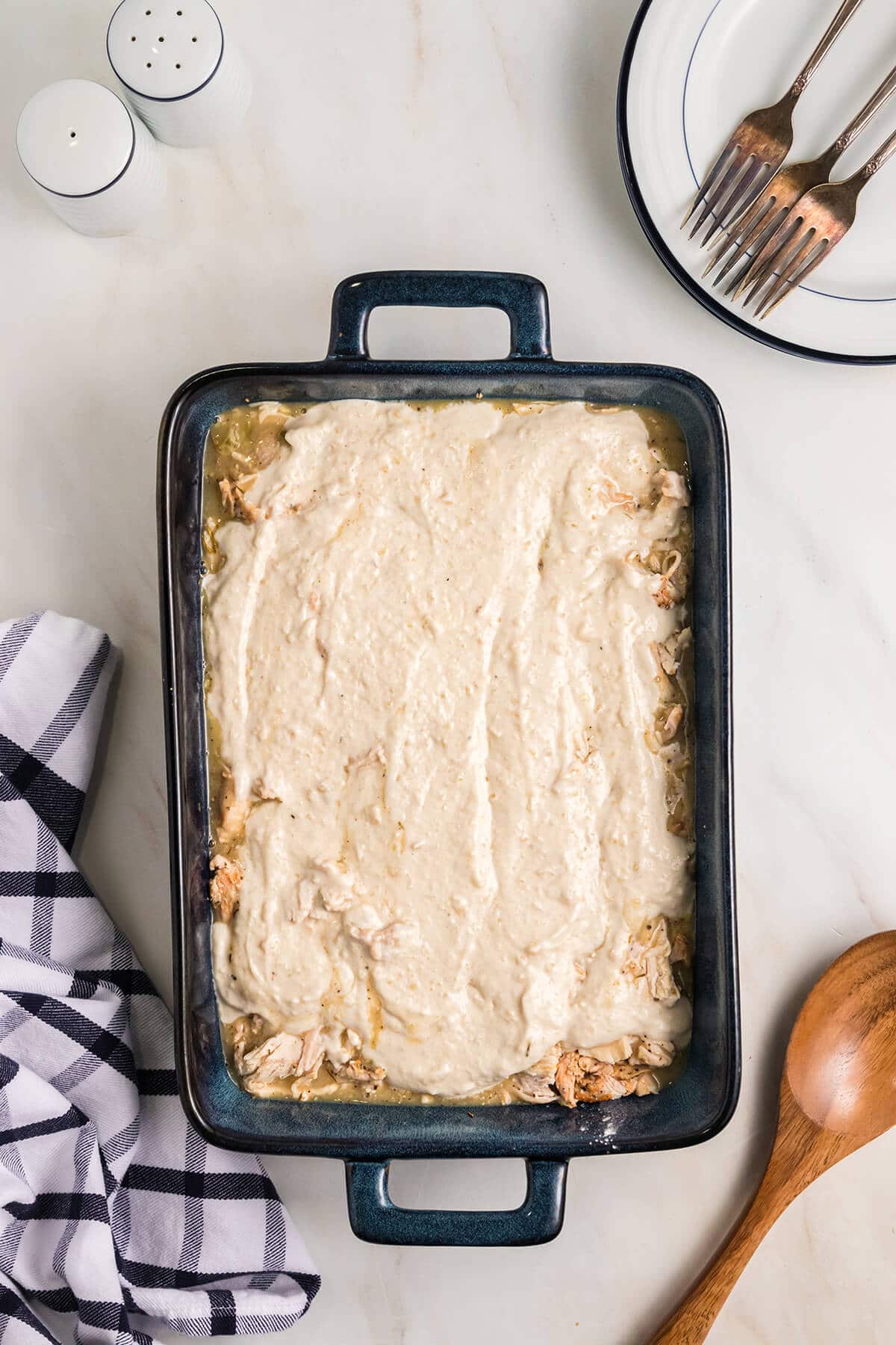Shredded chicken covered with cream of celery soup in a baking dish with a wooden spoon on the side.