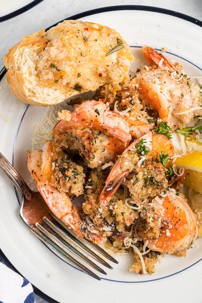 Closeup photo of a white plate with a serving of baked shrimp scampi with a fork and piece of bread.
