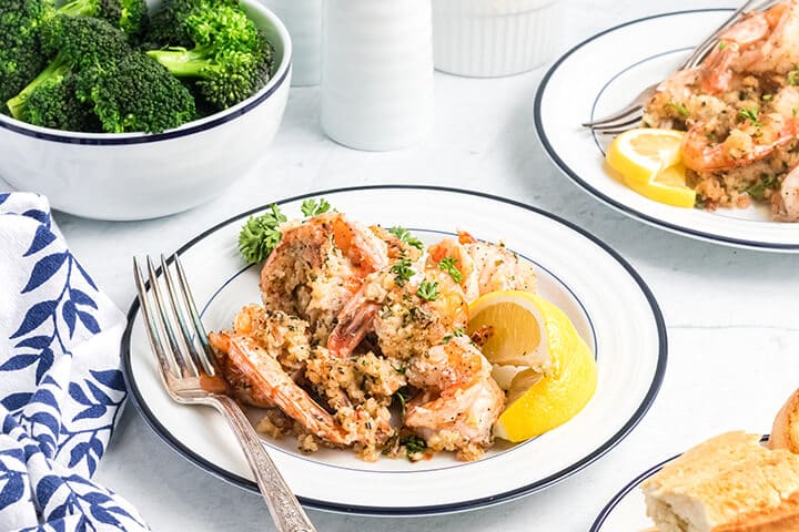 A white plate with a serving of baked shrimp scampi and a bowl of broccoli in the background.