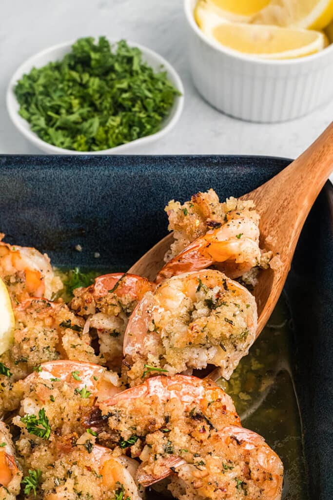 A wooden spoon scooping out baked shrimp scampi to serve.